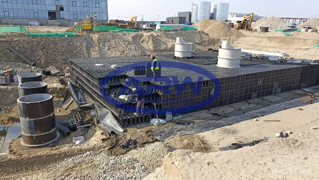 European project site installing an underground geocellular tank—stacked black crates on a compacted base with inspection risers and liner/geotextile being placed.