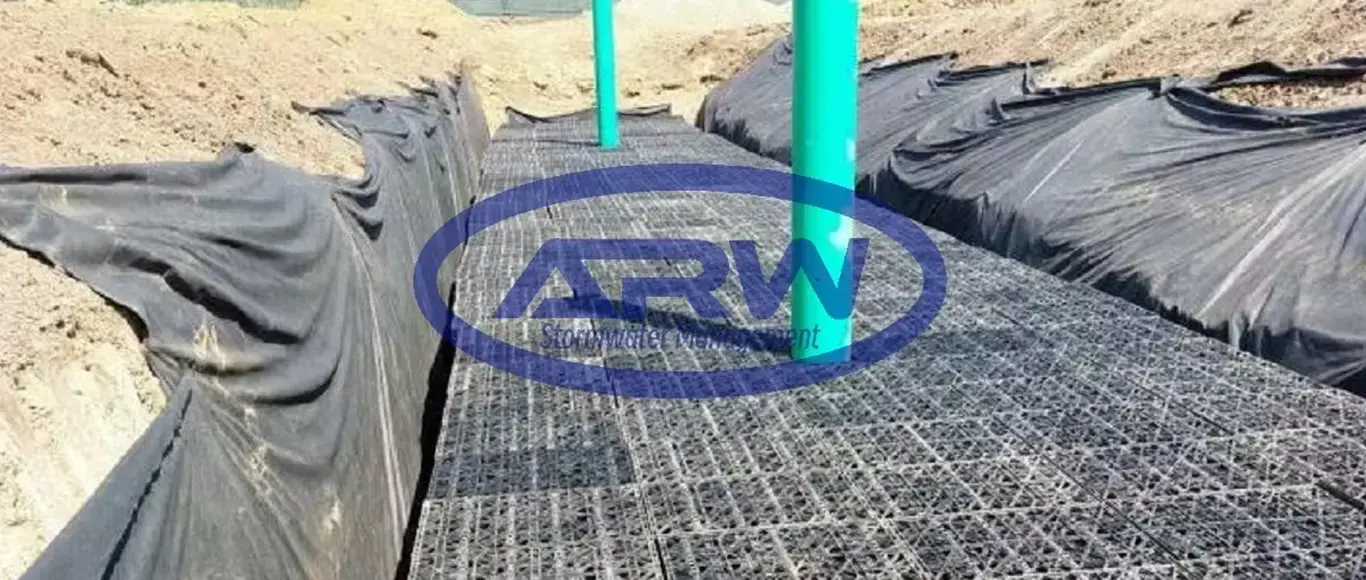 Worker installing a geocellular underground stormwater detention system at a US jobsite — plastic modules placed on a stone base in a geomembrane-lined excavation with geotextile protection.